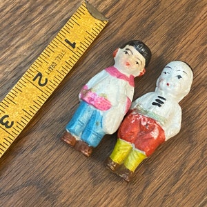 May include: Two vintage porcelain figurines of people wearing traditional clothing. One figurine is wearing a blue and white outfit with a pink flower design. The other figurine is wearing a red and yellow outfit with black stripes.