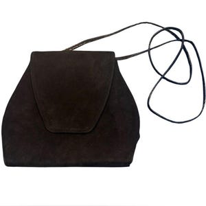 May include: A dark brown suede shoulder bag with a structured, trapezoidal silhouette and a large, pointed flap closure. The bag features a thin, matching dark brown shoulder strap that extends from the top sides, creating a clean, minimalist look.