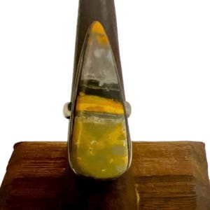 May include: A silver ring featuring a large, teardrop-shaped stone. The stone displays a unique pattern of yellow, gray, and black hues. The ring is set on a wooden display stand, showcasing the jewelry's design and craftsmanship.
