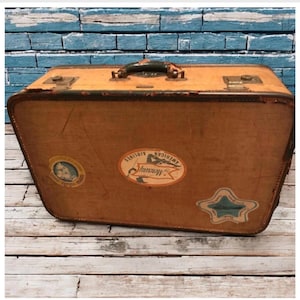May include: Vintage brown suitcase with a black handle and metal clasps. The suitcase has several stickers, including one that reads "American Airlines Mercury". The suitcase is on a wooden surface.