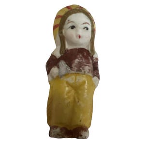 Puede incluir: Una peque&ntilde;a figurita de porcelana de una ni&ntilde;a que lleva un vestido amarillo, una camisa marr&oacute;n y un sombrero amarillo y rojo.