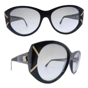 May include: Black oversized sunglasses with a cat-eye shape. The frames have gold accents on the corners and temples. The lenses are a gradient gray color. The sunglasses are shown from two different angles.
