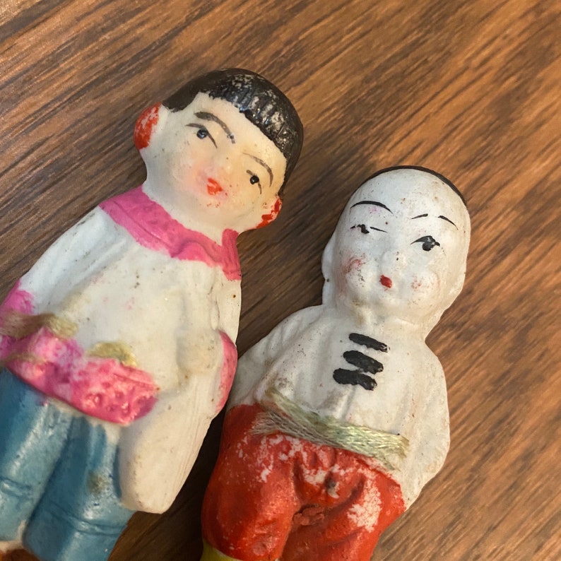 May include: Two porcelain dolls with painted faces and black hair. The doll on the left is wearing a white and pink outfit with blue pants. The doll on the right is wearing a white and red outfit with a black belt.