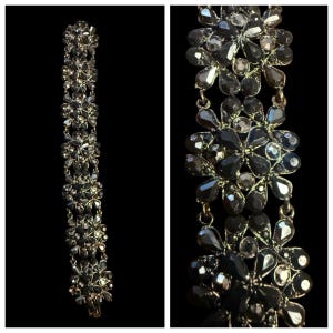 May include: A dark gray bracelet with a floral design. The bracelet features multiple flower-shaped clusters connected by small links. Each flower is made of dark gray and clear crystals, creating a sparkling effect. The background is black.
