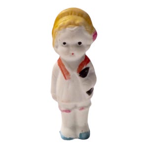Puede incluir: Una pequeña muñeca de porcelana con cabello rubio, una flor rosa y una bufanda roja y negra. La muñeca lleva un vestido blanco con detalles en rosa y azul.