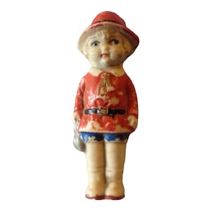 Puede incluir: Una muñeca de porcelana con un sombrero rojo, ropa roja y azul y una pequeña bolsa. La muñeca tiene una cara pintada con ojos azules y una boca roja.