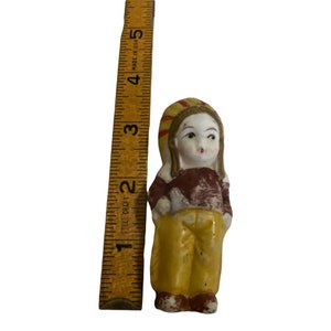 Puede incluir: Una peque&ntilde;a figurita de porcelana de una ni&ntilde;a que lleva un sombrero amarillo y rojo, una camisa marr&oacute;n y pantalones amarillos. La figurita mide aproximadamente 7,5 cm de alto.