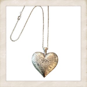 Peut inclure: Un collier avec un pendentif en forme de cœur argenté. Le pendentif en forme de cœur présente un motif floral en relief. Le collier a une chaîne délicate et une fermeture à fermoir. Le pendentif mesure environ 5 cm de large.
