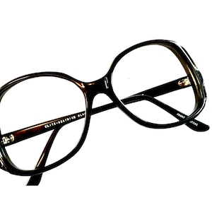 May include: Brown plastic framed glasses with a large, rounded lens shape. The glasses have the text "ELITE-BEATRICE BLACK" on the left arm and "FRANK JEWNE" on the right arm.