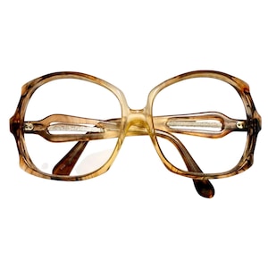 May include: Oversized eyeglasses with a translucent amber and brown tortoiseshell pattern frame. The temples feature a horizontal cutout design with a subtle silver glitter inlay. The frames have a rounded, wide shape with clear lenses.
