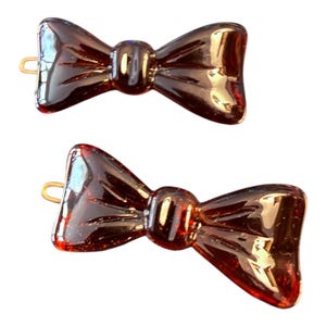 Peut inclure: Deux barrettes en forme de nœud papillon de couleur ambre foncé. Chaque barrette est dotée d'un fermoir en métal doré. Les nœuds ont une surface brillante et réfléchissante, et sont placés sur un fond blanc. Ces accessoires conviennent à diverses coiffures.