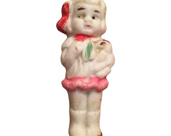 May include: A porcelain figurine of a girl with a red bow in her hair, wearing a pink dress and holding a small white doll.