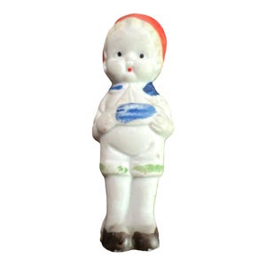 Puede incluir: Una pequeña figurita de porcelana de un niño que lleva una camisa blanca, un cuello a rayas azul y blanco, pantalones verdes y un sombrero rojo. La figurita tiene una cara pintada con ojos azules, labios rojos y una tez blanca.