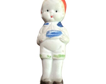 May include: A small porcelain figurine of a boy wearing a white shirt, blue and white striped collar, green pants, and a red hat. The figurine has a painted face with blue eyes, red lips, and a white complexion.