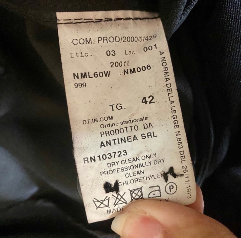 May include: A white tag with black text attached to a black garment. The tag includes the text "COM. PROD/2000/01429", "Etic. 03 Lav. 001", "NML60W 20011 NM006 999", "TG. 42", "DT.IN.COM Ordine stagionale PRODOTTO DA ANTINEA SRL RN 103723 DRY CLEAN ONLY PROFESSIONALLY DRY CLEAN CHLORETHYLEN MADE", and "A NORMA DELLA LEGGE N.883 DEL 26/11/1973".