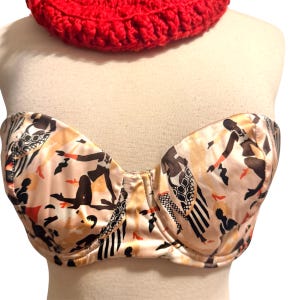 May include: A strapless bra with a beige background and a pattern of black and brown figures. The bra is on a mannequin. A red crocheted item is above the bra.