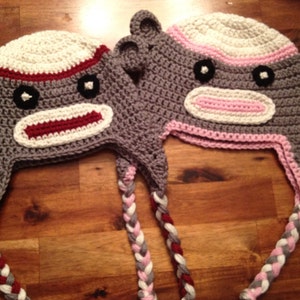 May include: Two crocheted monkey hats with long tails. One hat is gray with white and burgundy stripes, the other is gray with white and pink stripes.