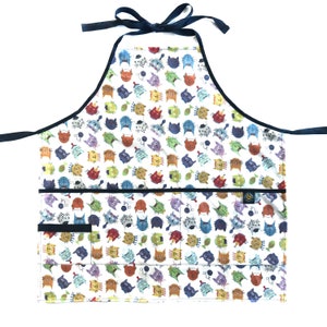 May include: A white apron with a colorful cat-themed pattern. The apron has a dark blue trim and ties, with two front pockets. The cat faces are in various colors, including orange, green, and purple.