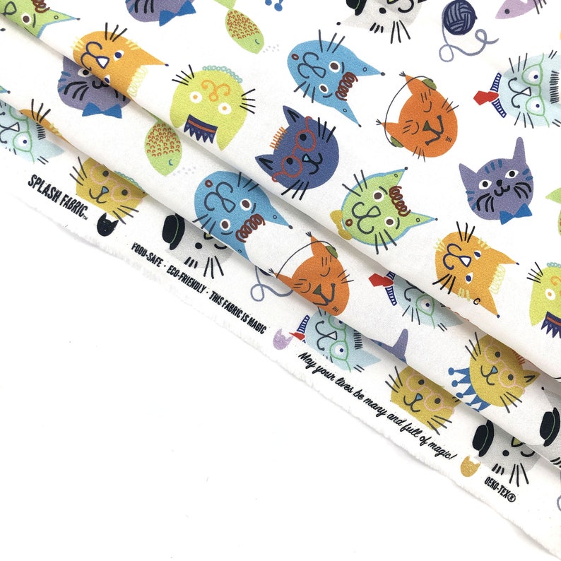 May include: White fabric with a repeating pattern of colorful cartoon cats. The cats have various expressions and wear accessories like glasses and bow ties. The fabric also features text that reads "SPLASH FABRIC" and "FOOD-SAFE - ECO-FRIENDLY - THIS FABRIC IS MAGIC."