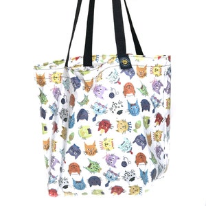 May include: White tote bag with a colorful cat face pattern. The bag has black handles and a tag with text. The cat faces are in various colors, including orange, blue, and green. The bag is made of fabric.