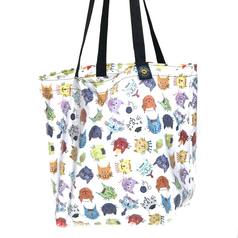 May include: White tote bag with a colorful cat face pattern. The bag has black handles and a tag with text. The cat faces are in various colors, including orange, blue, and green. The bag is made of fabric.