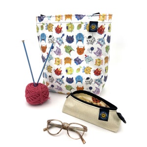 May include: A white tote bag with a colorful cat pattern, a pink yarn ball, blue knitting needles, a beige pencil case, and eyeglasses. The tote bag and pencil case have a circular logo. The eyeglasses have a tortoise shell pattern.