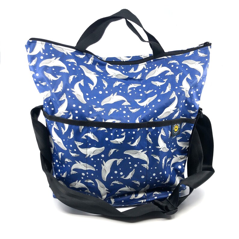 May include: A blue tote bag with a white whale pattern and white polka dots. The bag has a black zipper and a black shoulder strap.
