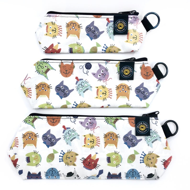 May include: Three white pencil cases with a colorful cat pattern. Each case has a black zipper, a black loop, and a small tag that says "SPLASH FABRIC". The cat illustrations are in various colors and styles.