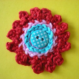 May include: A crocheted flower with red, pink, and blue yarn, featuring a blue button with white polka dots in the center.