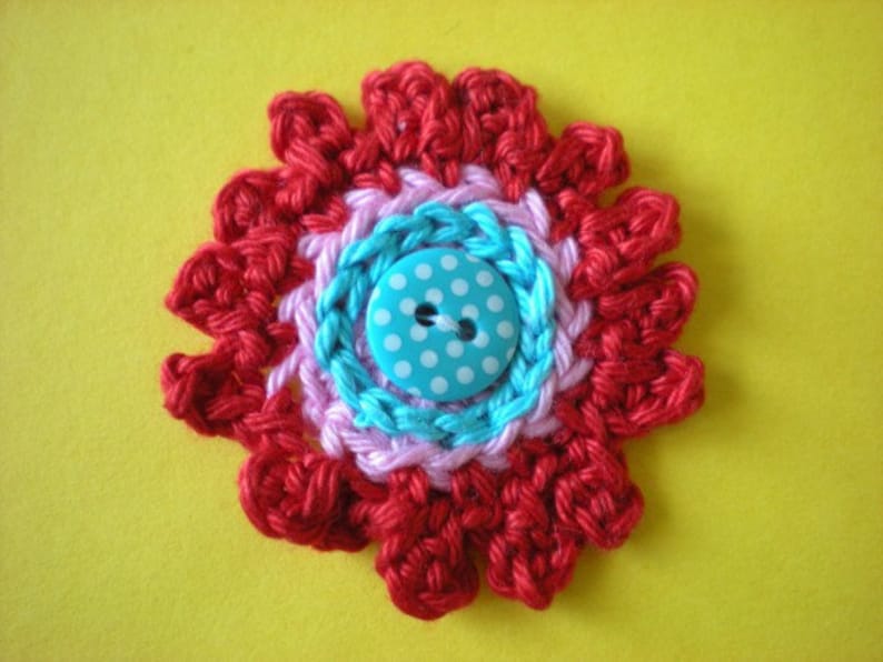 May include: A crocheted flower with red, pink, and blue yarn, featuring a blue button with white polka dots in the center.