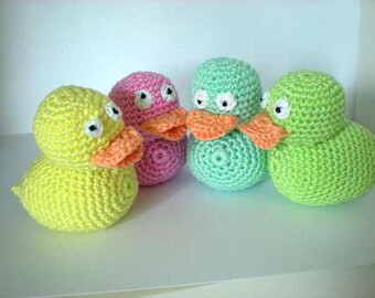 Op de afbeelding: Vier gehaakte rubberen eendjes in verschillende kleuren: geel, roze, groen en lichtgroen. Elk eendje heeft witte ogen en een oranje snavel.