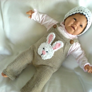 May include: A baby doll wearing a knitted brown jumpsuit with a white bunny applique and a white crocheted hat.