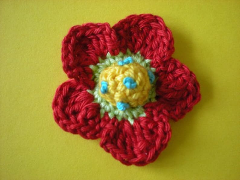 May include: A crocheted flower with red petals and a yellow center with blue accents.