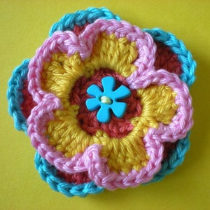 May include: A crocheted flower with layers of pink, yellow, and red yarn. The center of the flower has a blue button with a five-petal design.