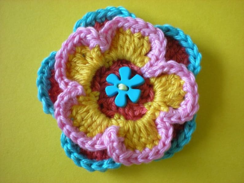 May include: A crocheted flower with layers of pink, yellow, and red yarn. The center of the flower has a blue button with a five-petal design.