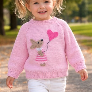 Puede incluir: Una niña pequeña con un suéter rosa tejido con un aplique de perro y globo de corazón. El suéter se combina con pantalones blancos. La niña sonríe y tiene coletas rubias.