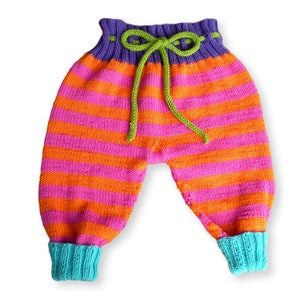 May include: Striped knitted baby pants with a purple waistband and green drawstring. The pants have orange and pink stripes and turquoise cuffs.