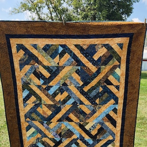 May include: A quilt with a geometric pattern of blue, brown, and tan fabric. The quilt is hanging on a clothesline outside.