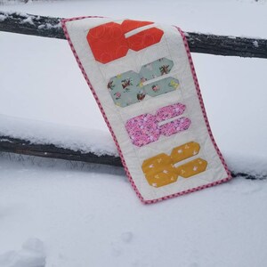 May include: A white fabric banner with a red and white checkered border. The banner has the word "SIX" spelled out in large, colorful letters. The letters are made of different patterned fabrics. The banner is hanging on a wooden fence post in a snowy landscape.