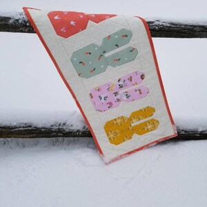 May include: A white quilted table runner with an orange border features a patchwork design of bunnies with different patterns. The runner is hanging on a wooden fence post in a snowy setting.