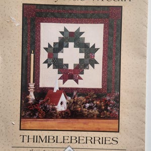 May include: A printed pattern titled "Countryside Wreath" by Thimbleberries. The design features a quilted wreath with star-shaped elements in shades of green and red. A small church and candle are also depicted.