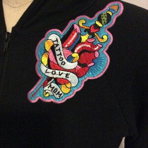 May include: Black zip-up hoodie with a colorful embroidered patch. The patch features a heart, dagger, and banner with the words "Tattoo Love Mom". The design is outlined in pink and blue, with yellow and red accents.