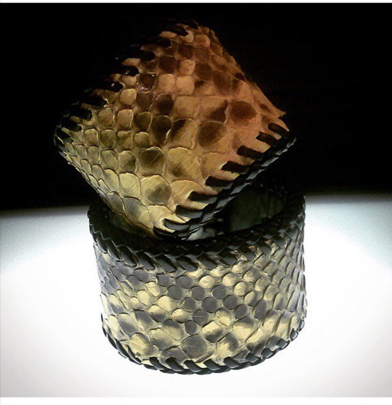 K&ouml;nnte beinhalten: Zwei Schmuckst&uuml;cke, ein Armband und ein Ring, aus einem schlangenlederartigen Material. Das Armband ist ein breites Band, und der Ring ist ein kleineres, quadratisch geformtes St&uuml;ck. Beide haben einen schwarzen, gen&auml;hten Besatz.