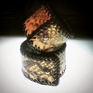 K&ouml;nnte beinhalten: Zwei gestapelte Lederarmb&auml;nder mit Schlangenmuster in Beige, Braun und Schwarz. Jedes Armband hat eine schwarze geflochtene Ledereinfassung und einen silbernen Knopfverschluss. Die Armb&auml;nder sind handgefertigt und auf einer wei&szlig;en Oberfl&auml;che ausgestellt.