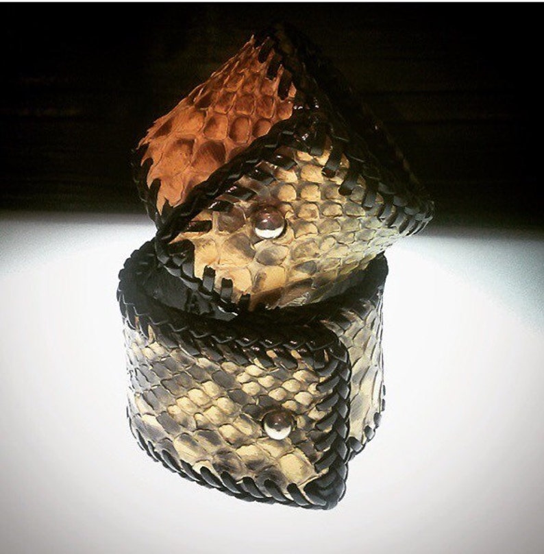 K&ouml;nnte beinhalten: Zwei gestapelte Lederarmb&auml;nder mit Schlangenmuster in Beige, Braun und Schwarz. Jedes Armband hat eine schwarze geflochtene Ledereinfassung und einen silbernen Knopfverschluss. Die Armb&auml;nder sind handgefertigt und auf einer wei&szlig;en Oberfl&auml;che ausgestellt.