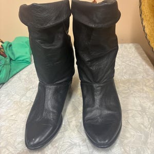 May include: A pair of black leather boots with a folded-over cuff. The boots have a pointed toe and a low heel. The leather appears slightly distressed, giving them a vintage look. The boots are standing upright.