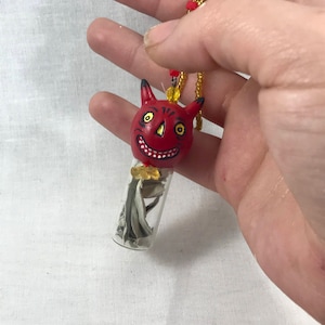 May include: A unique necklace featuring a red devil-faced pendant with yellow eyes and a smiling mouth. The pendant is attached to a small glass vial filled with dried herbs. The necklace has a gold beaded chain with red accents.