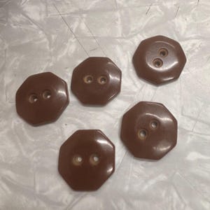 May include: Five brown, octagonal buttons with two or three holes each. The buttons have a glossy finish and are arranged on a wrinkled, light-colored surface. These buttons are suitable for sewing projects.