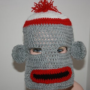 May include: A grey crocheted monkey hat with red and white stripes, a red mouth, and black eyes.