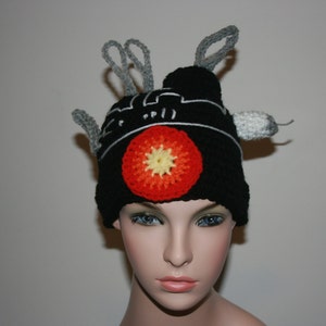 May include: A black knitted hat with a red and yellow circle design and gray crocheted antennas. The hat is worn by a person with a light complexion and blue eyes.